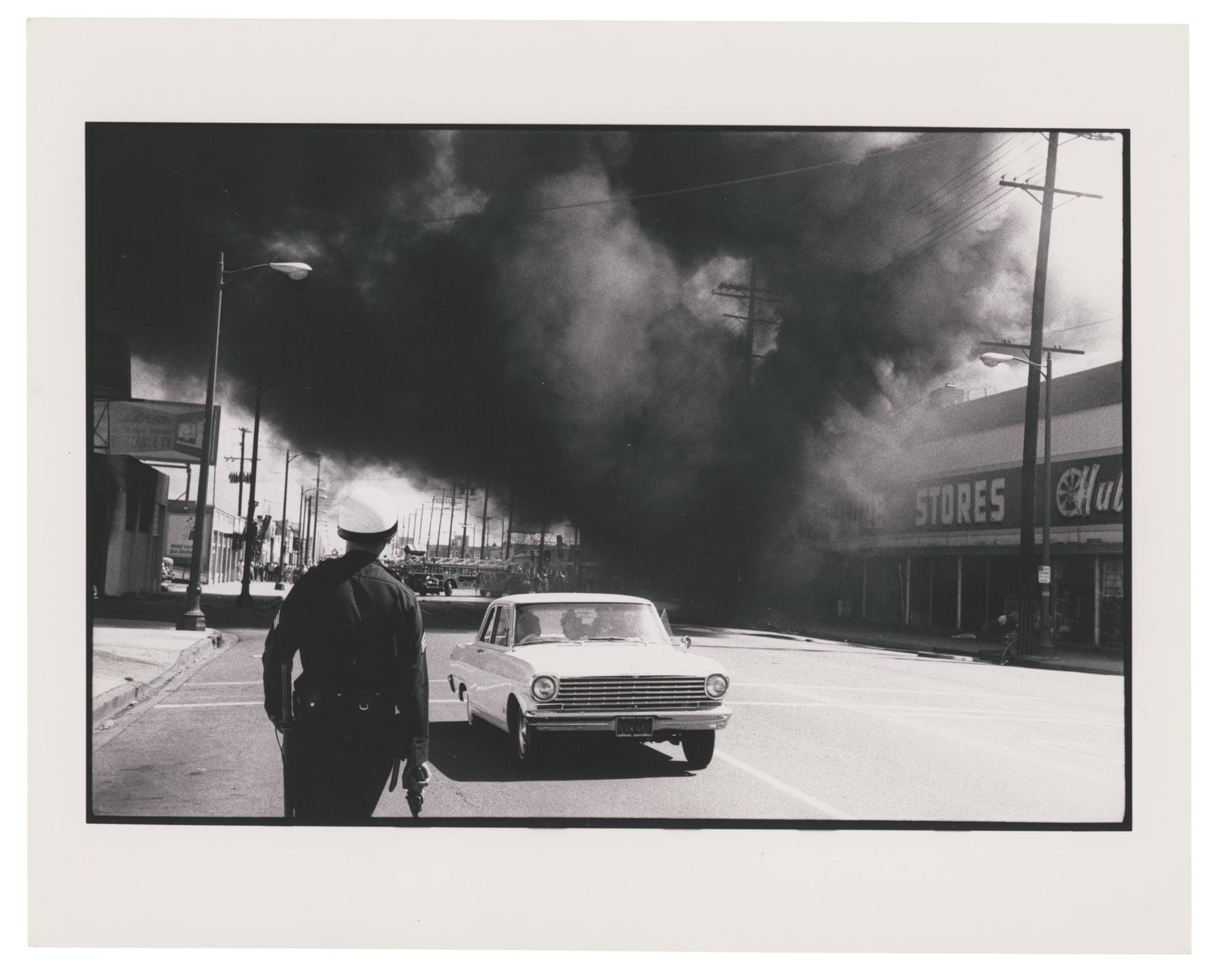 On a street, a plume of smoke comes out of a storefront. A helmeted police officer walks towards it with a gun, back to the camera, while a car drives away from it.