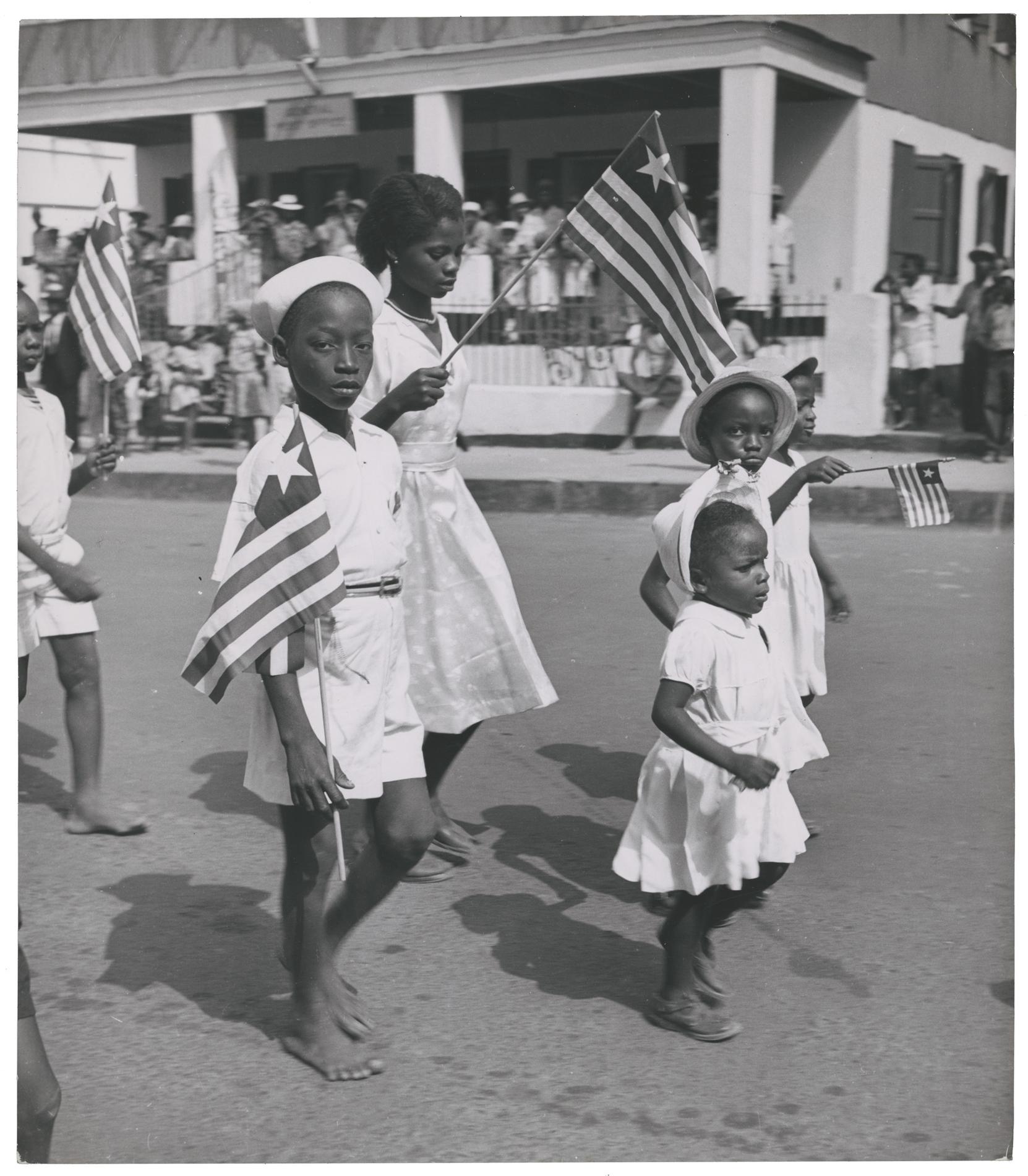 Two black little black boys and three little black girls wearing white while carrying Liberian flags and walking down the street.