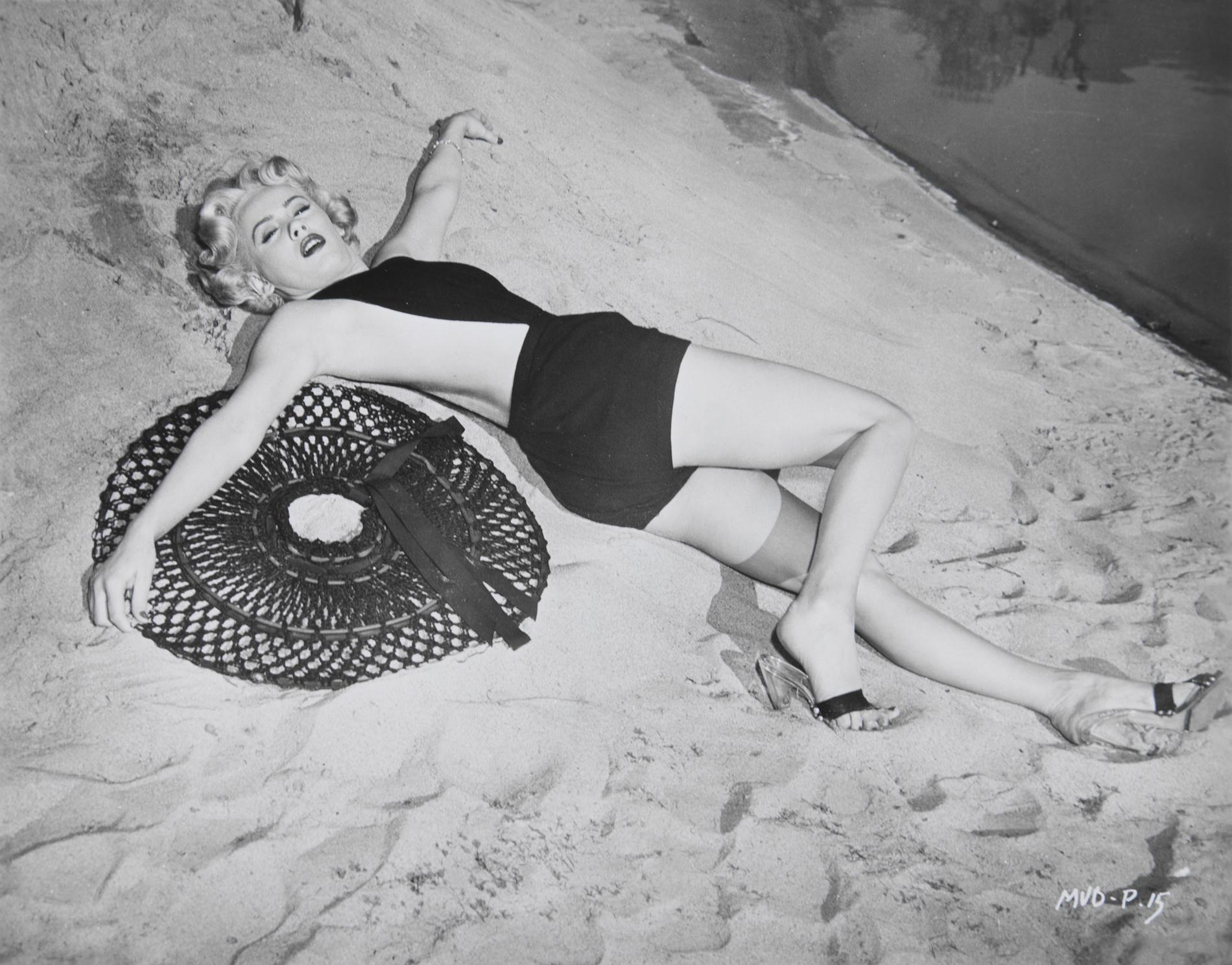 A blonde woman laying in the sand, arms outstretched, wearing a black swimsuit.