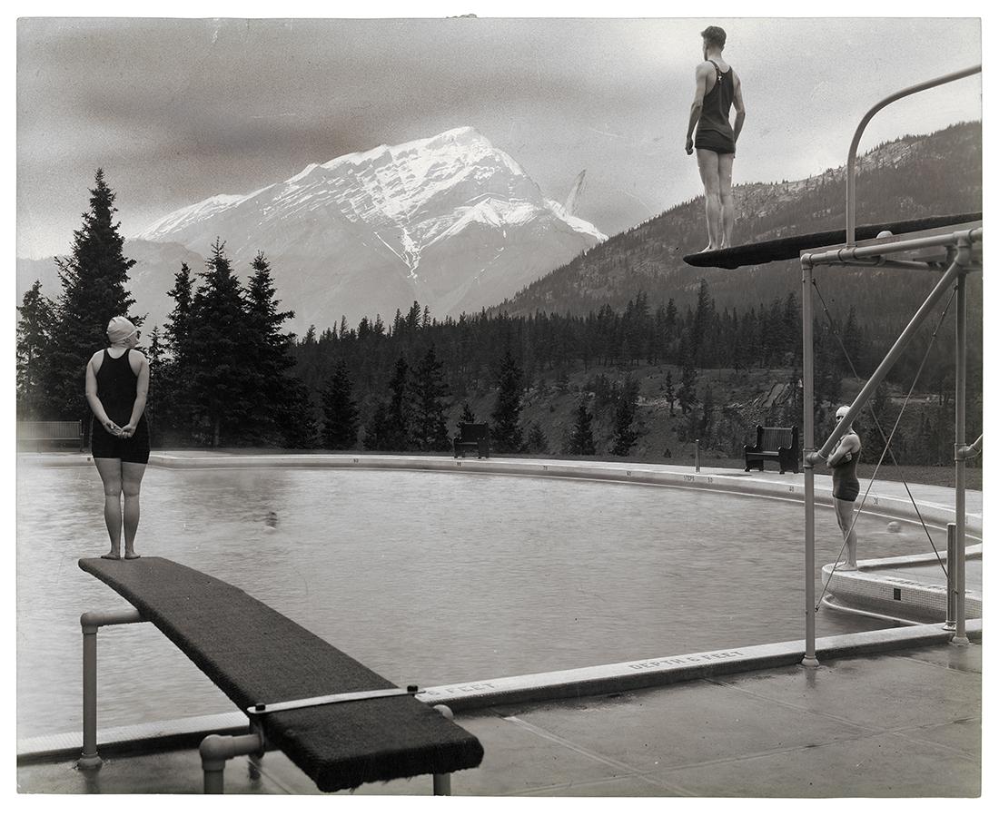 A black and white photograph of a man and a woman standing on two diving boards, with a view of a snowy mountain and forests in the background.