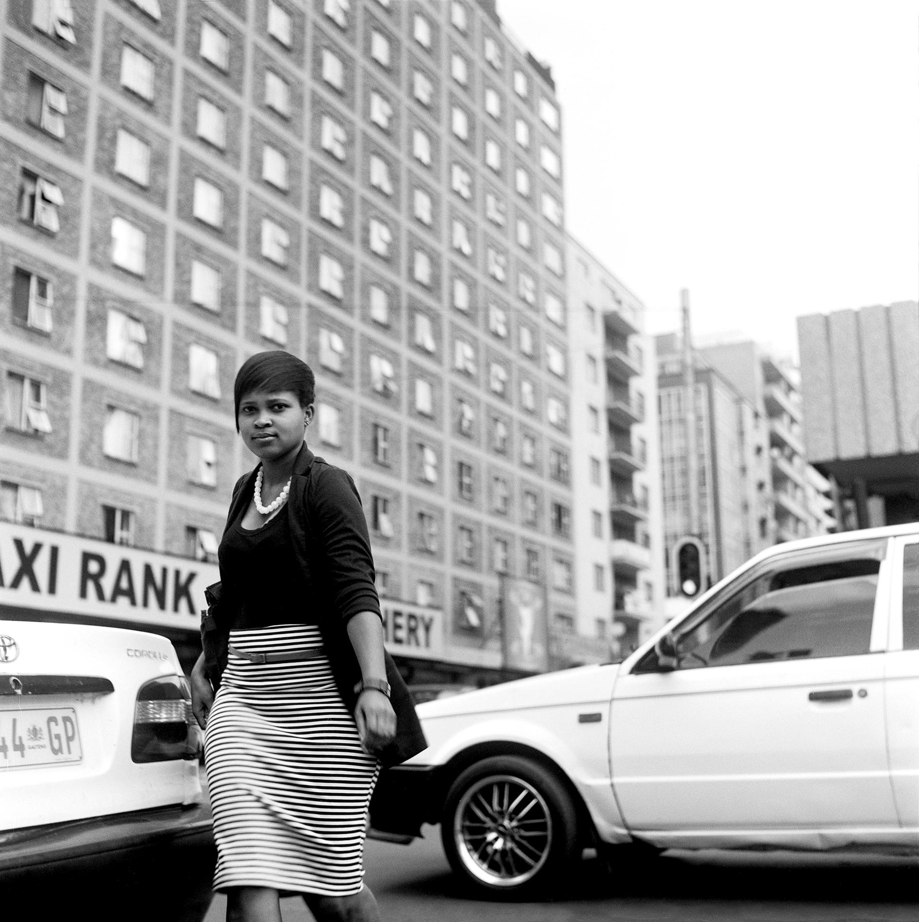 Black and white photo of a woman walking between cars amidst tall apartment buildings.