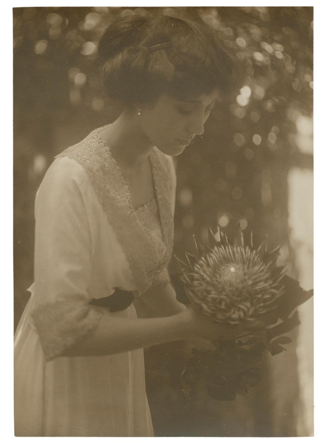 Woman in white dress holding a large flower. Black and white photograph by Minna Keene.