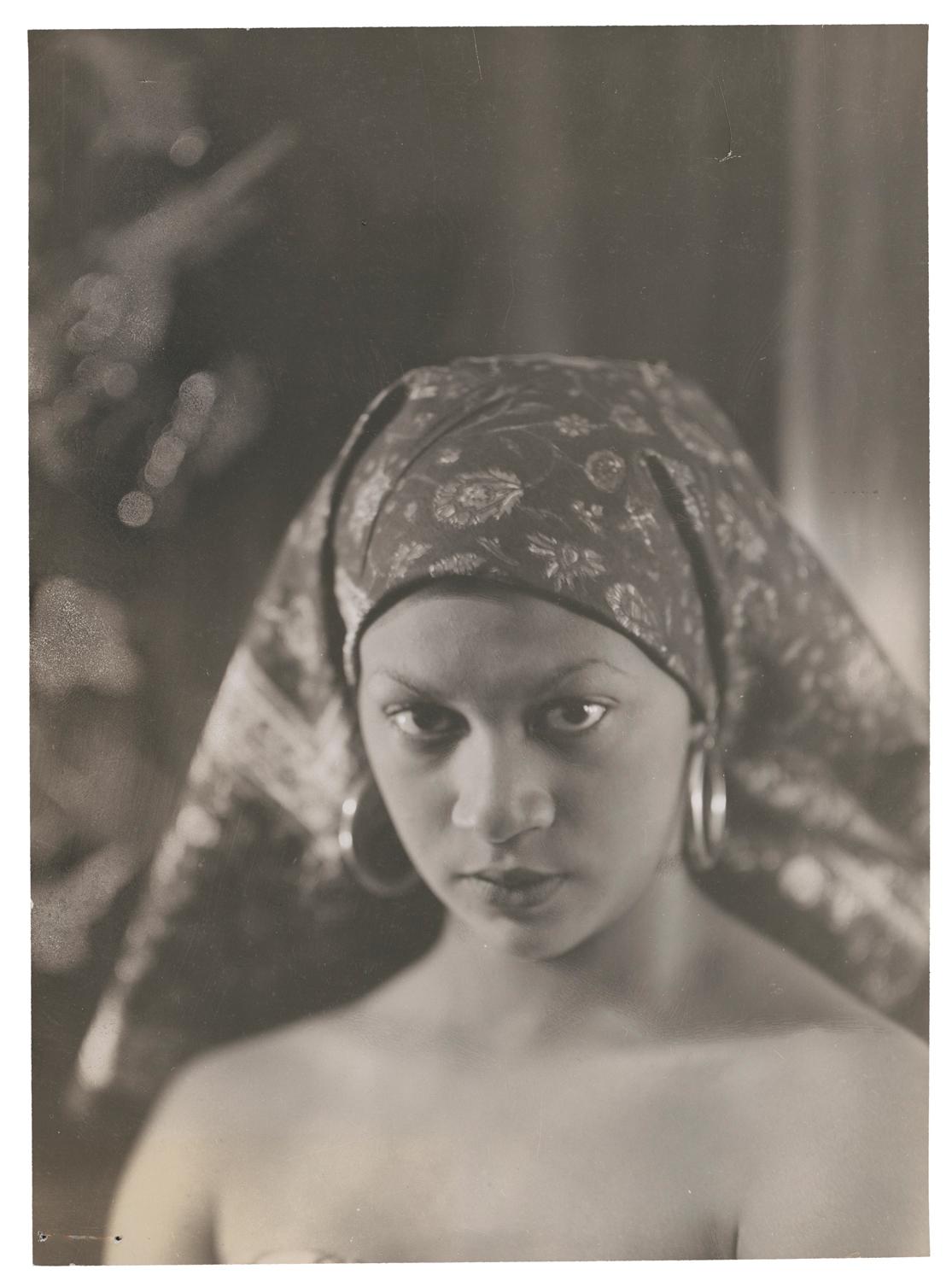 A woman wearing large hoop earrings and a headscarf looking directly into the camera. Black and white photograph by Violet Keene Perinchief.