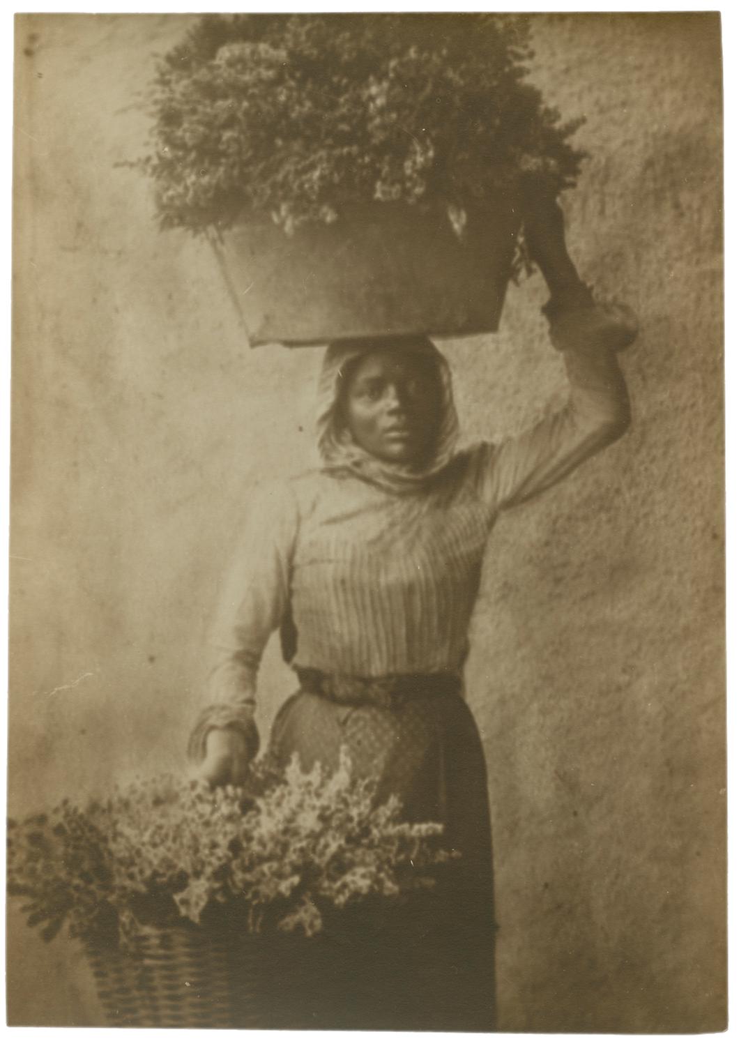 Portrait of a woman holding a large vessel with flowers on her head with her right hand and a large basket of flowers  in her left hand by her waist. She is looking into the camera. Black and white photograph by Minna Keene.