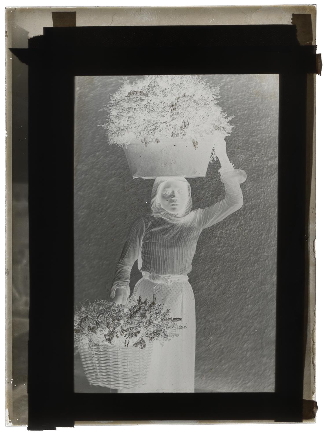 A negative of a portrait of a woman holding a large vessel with flowers on her head with her right hand and a large basket of flowers in her left hand by her waist. She is looking into the camera. Black and white photograph by Minna Keene.
