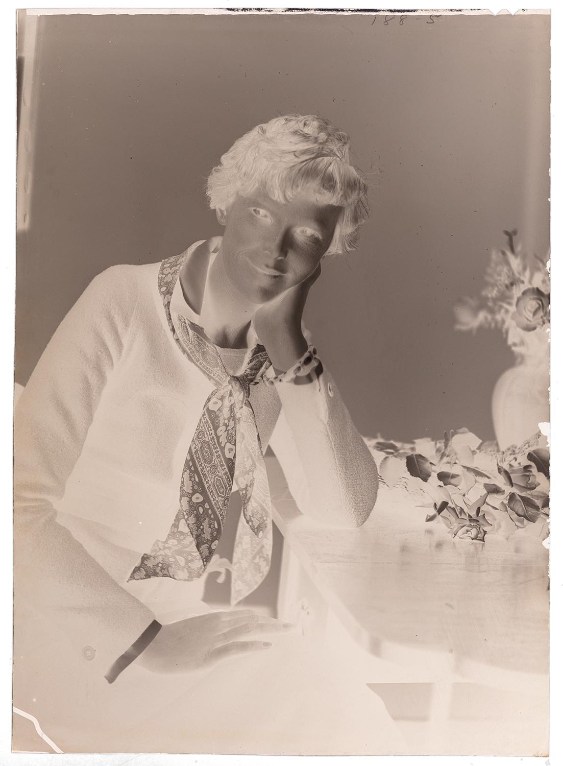 A negative portrait of a woman sitting down with her hand on her cheek and the other on her lap looking towards the camera. There is a vase of flowers beside her. Black and white photograph by Violet Keene Perinchief.