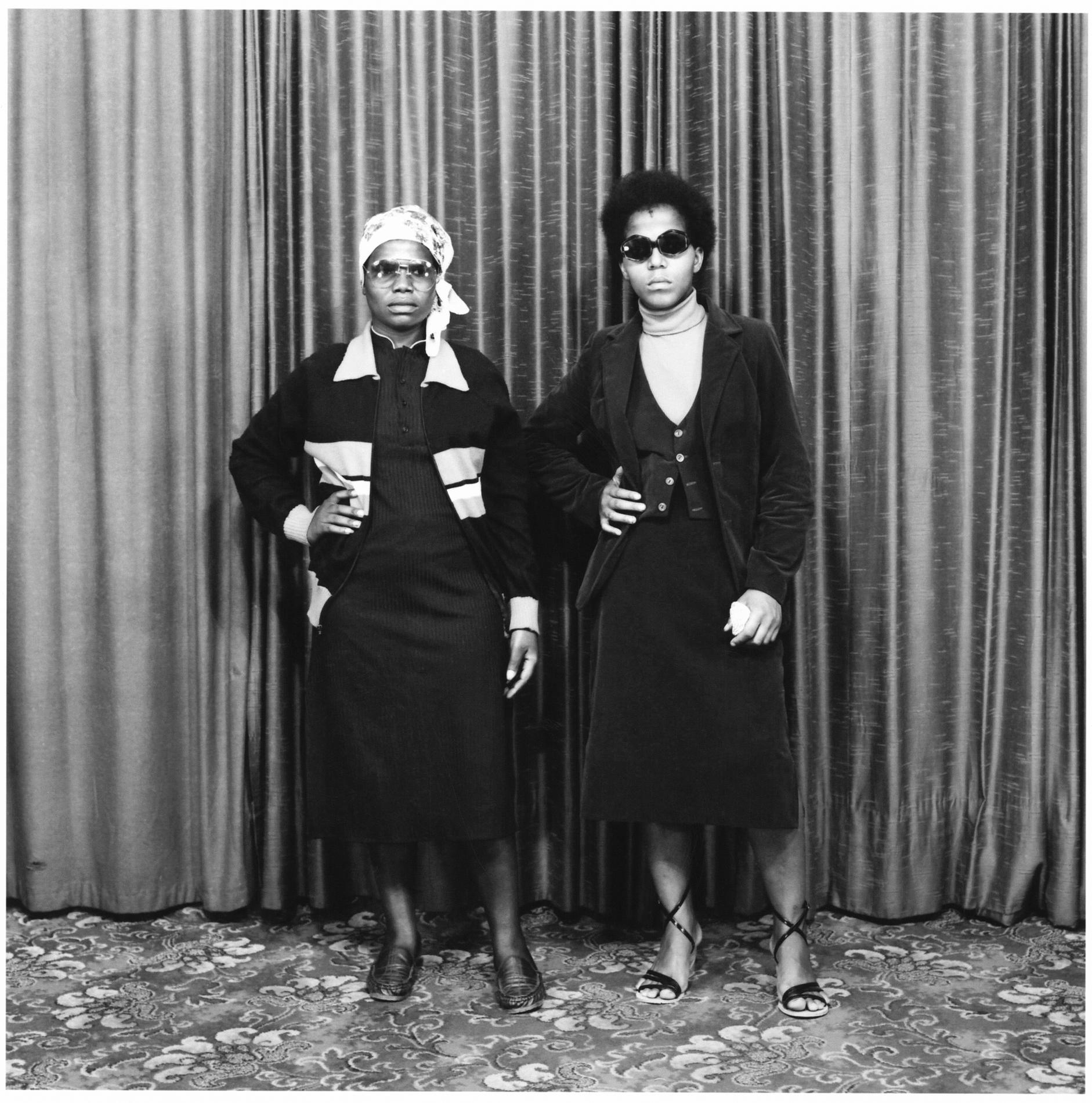 In this black and white image, two women stand confidently with their hands on their hips in front of a curtained backdrop wearing sunglasses