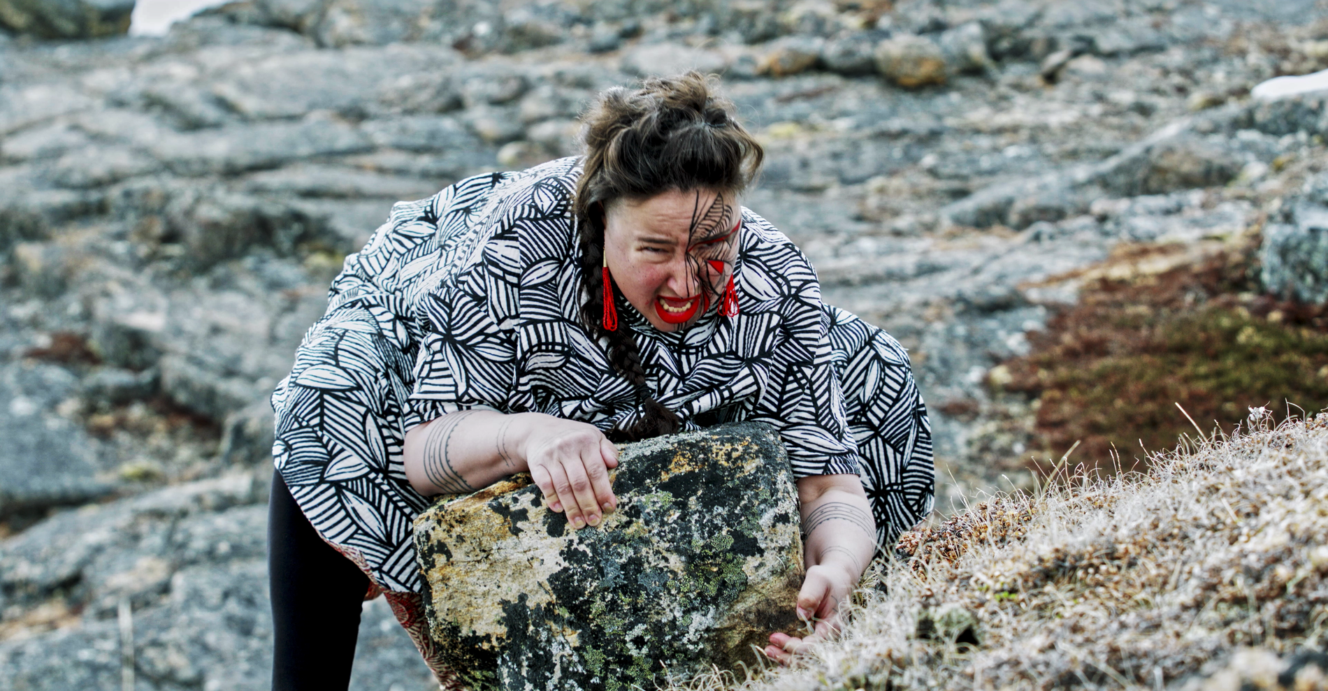 Indigenous woman in face paint crouches behind rock in plains landscape. 2000w