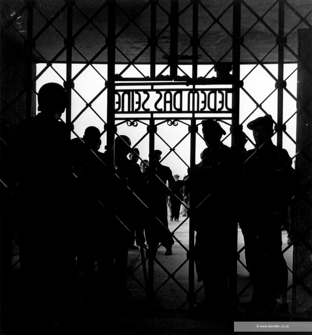 Black and white photo of gated entrance to concentration camp with the silhouettes of a crowd of people behind the bars.