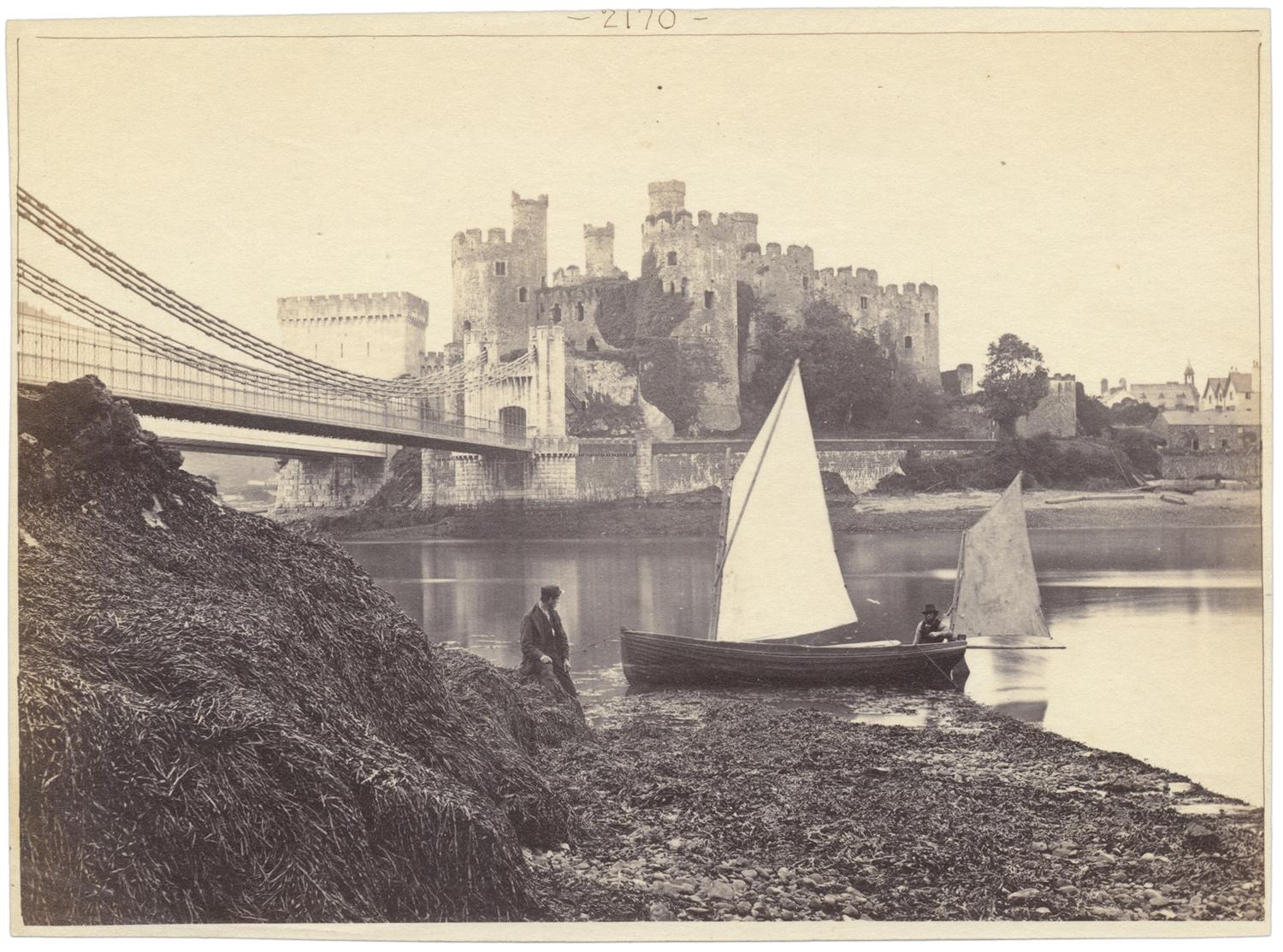 In the foreground, two sailboats near the shore, with a suspension bridge shown at the left leading to a castle.
