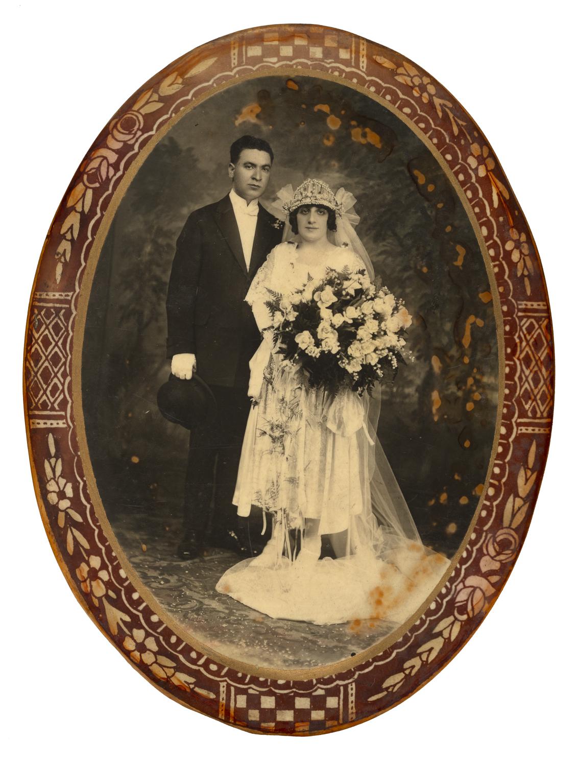 Couple stands posed for a wedding portrait. Woman bride wearing a crown carries large bouquet while male stands slightly behind her.