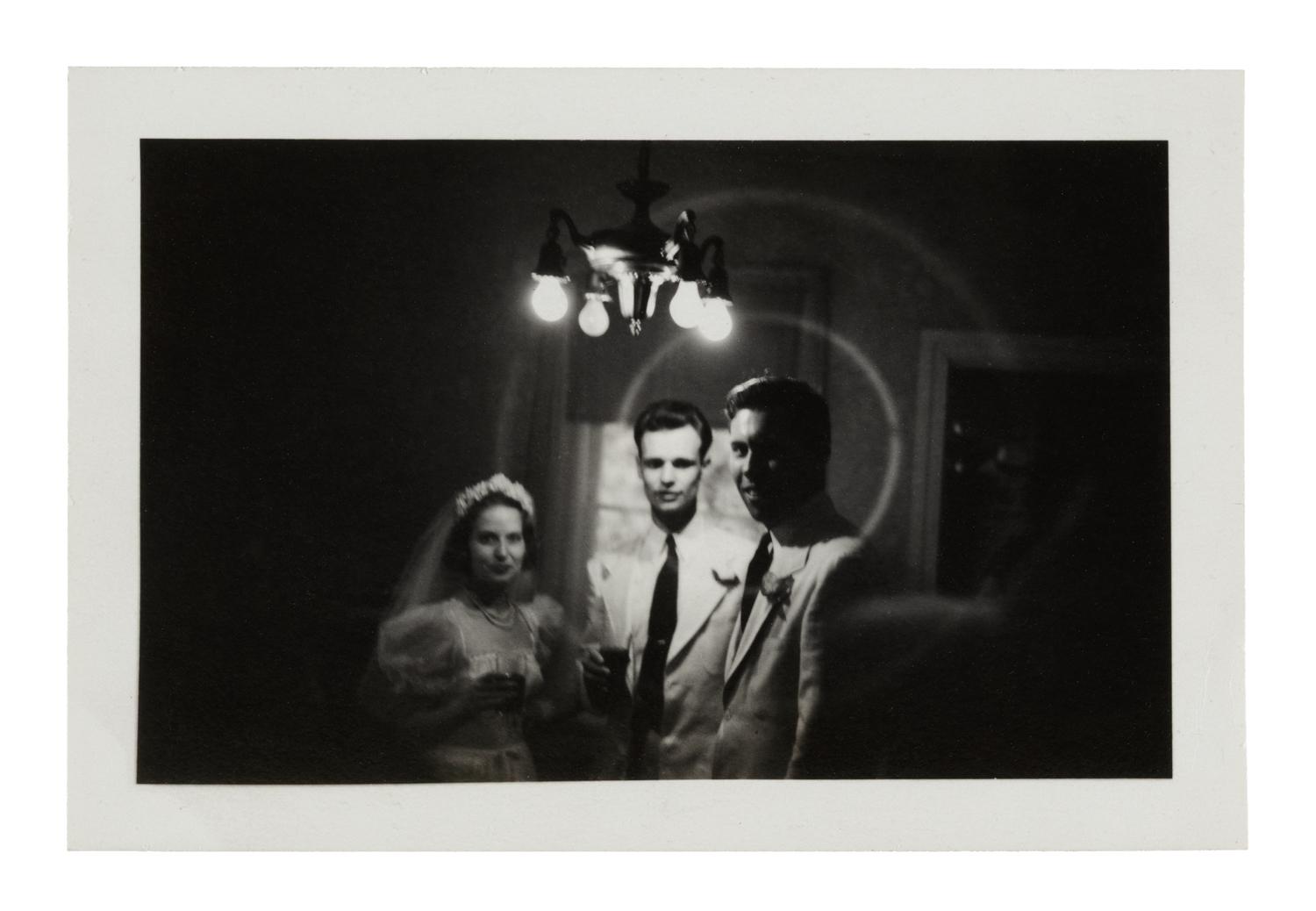 newlywed man and woman stand beside groomsman beneath bright overhead lightbulbs in a vignette of darkness