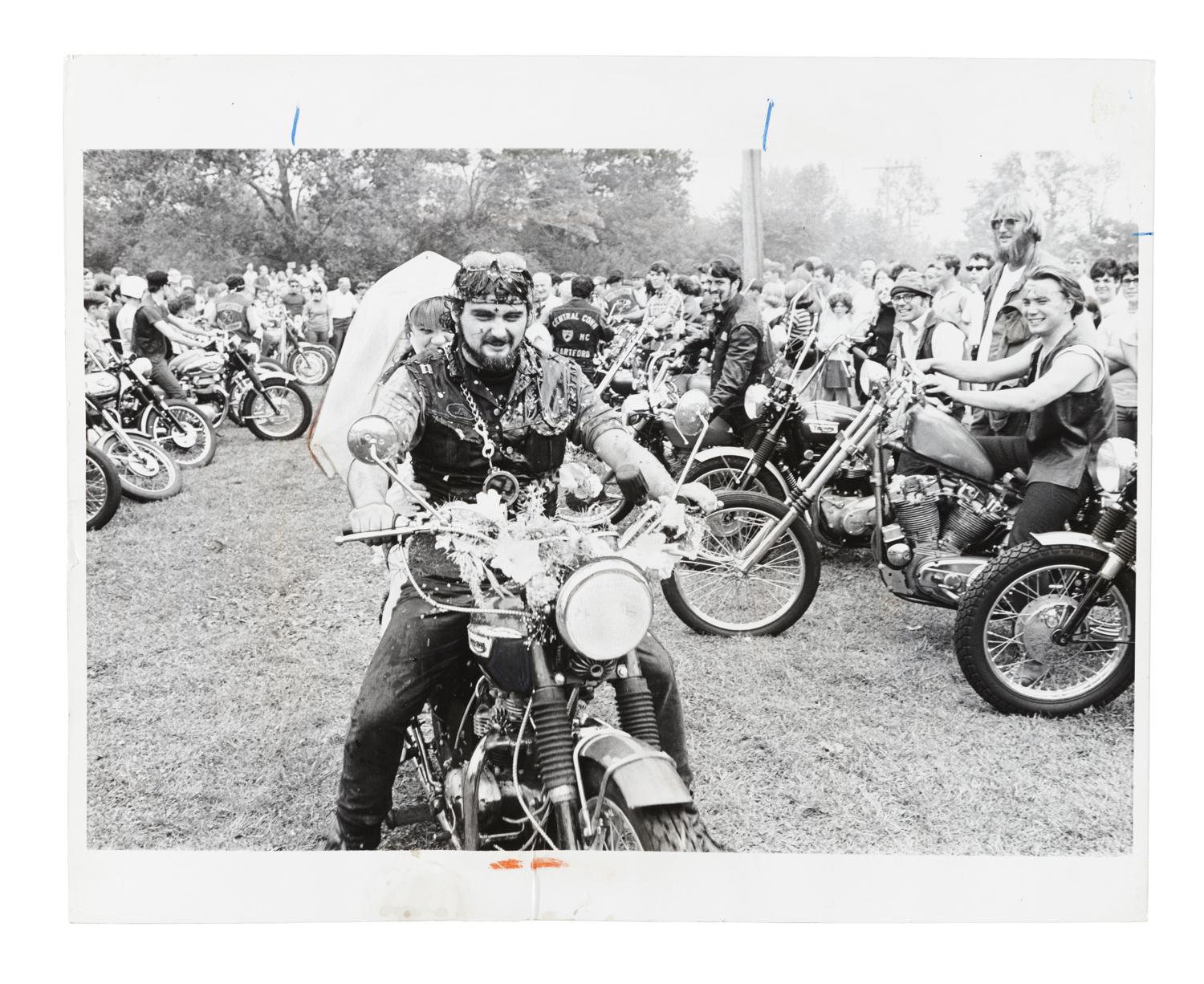 Man with leather vest on motorbike with veiled bride sat behind him. The pair are surrounded by other people on motorbikes.