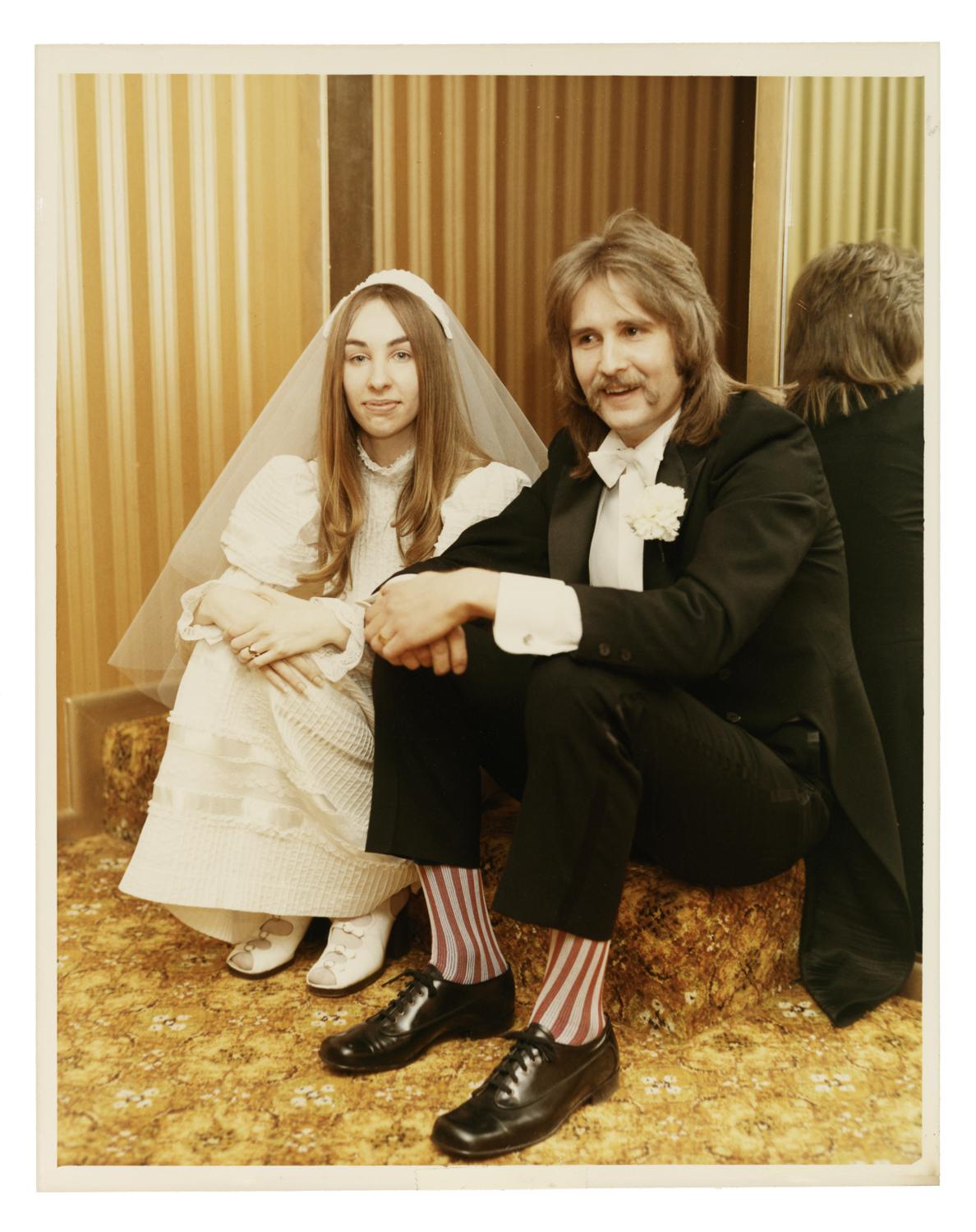newlywed bride and groom sit on a patterned carpet floor. Bride wears a veil, groom has a mustache, a mullet and red striped socks.