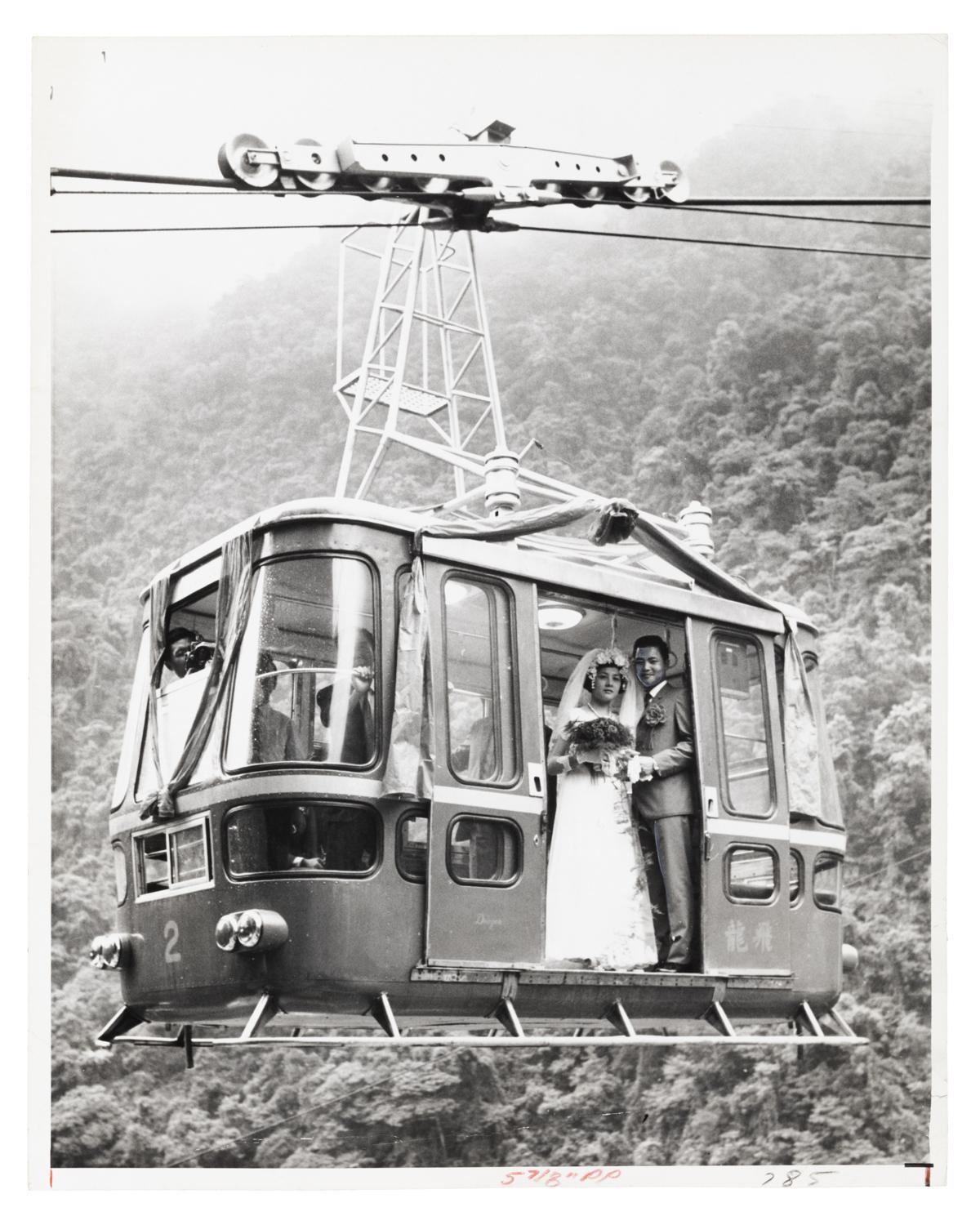 newly wed husband and wife stand in the open door of a cable car, shot from below in front of a mountain