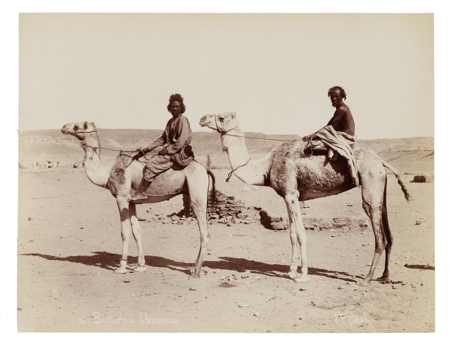 A sepia-toned photo showing two black men seated on two camels in a desert landscape. The person in the lead is wearing loose pants and top, while the person in the back is shirtless and draped in a loose fabric from the waist down.