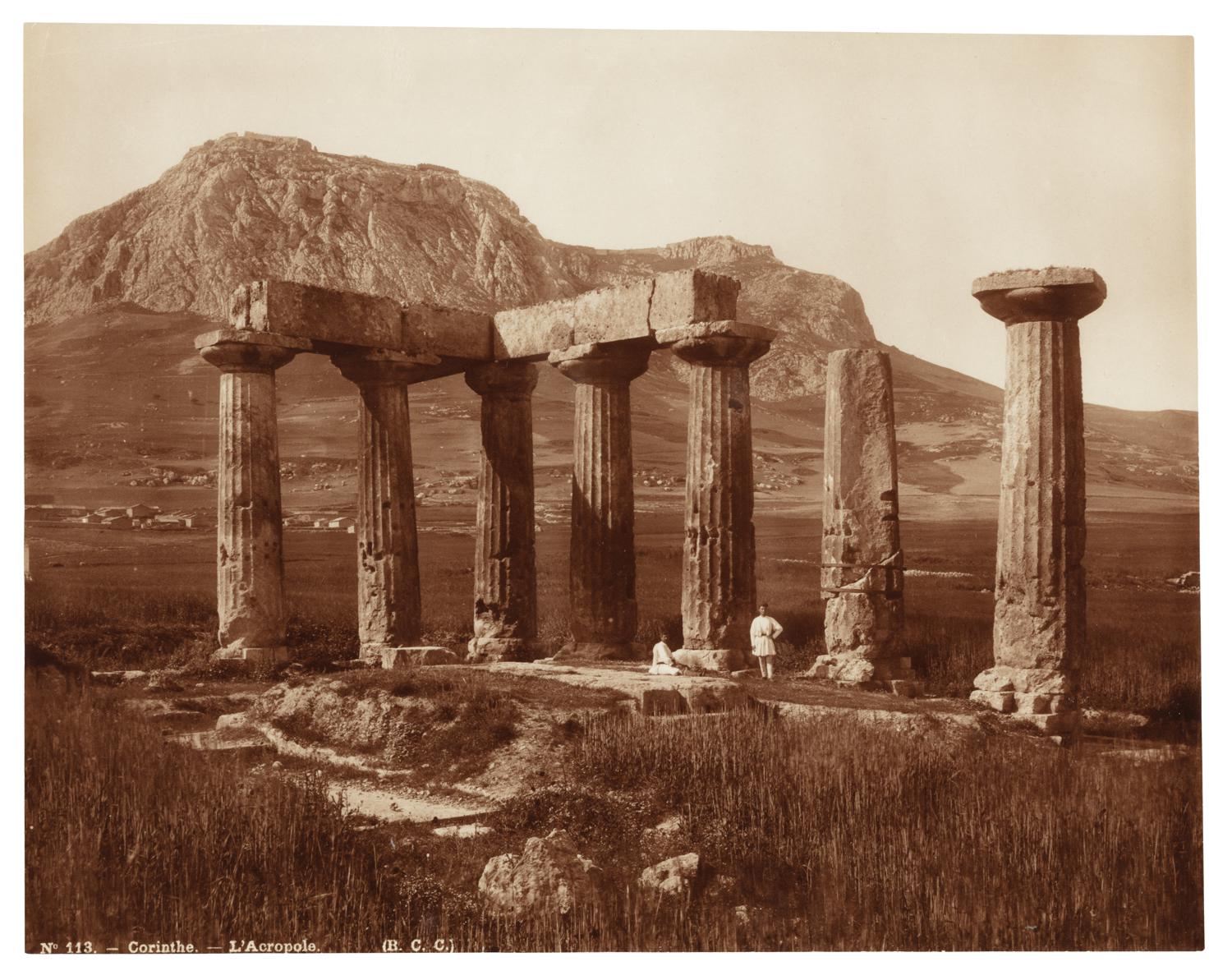 Two people in front of an acropolis in Corinth, Greece. The acropolis is located in a grassy field, with a mountain in the background.