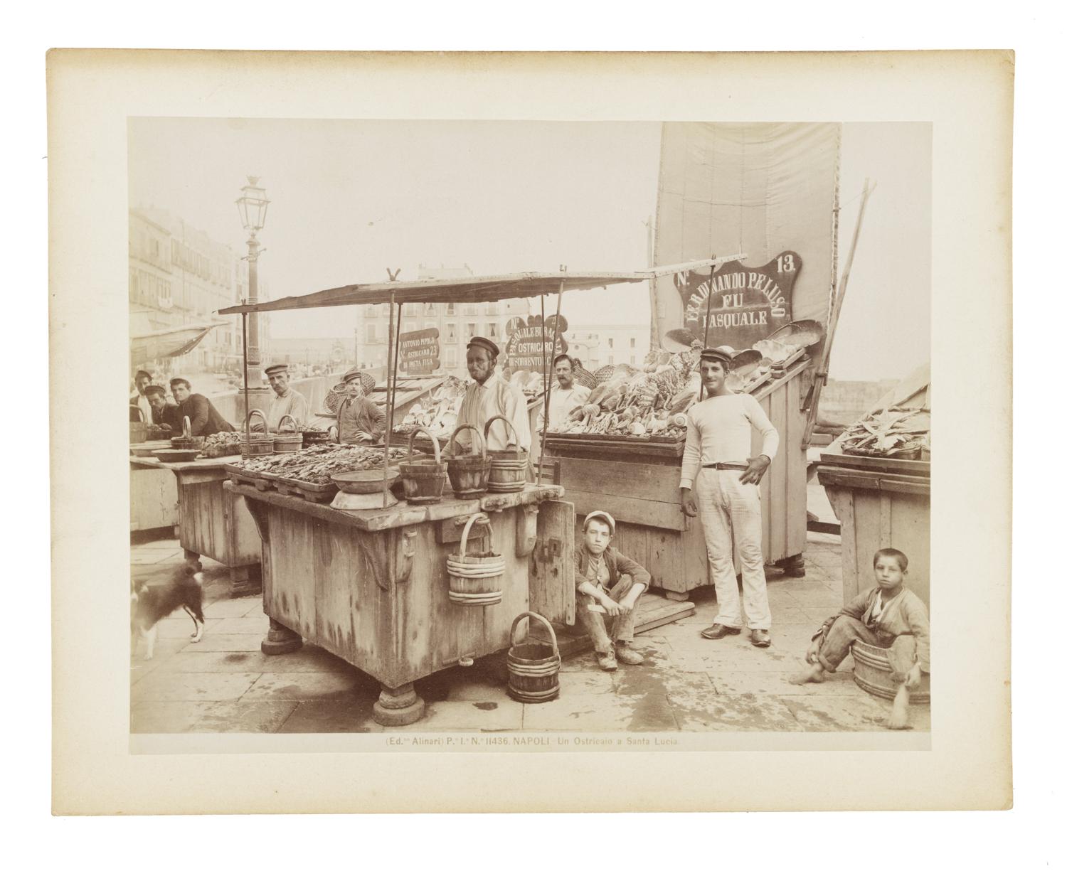 An oyster merchant pictured at a booth showcasing his wares. A number of other men and boys are pictured around the booth looking into the camera