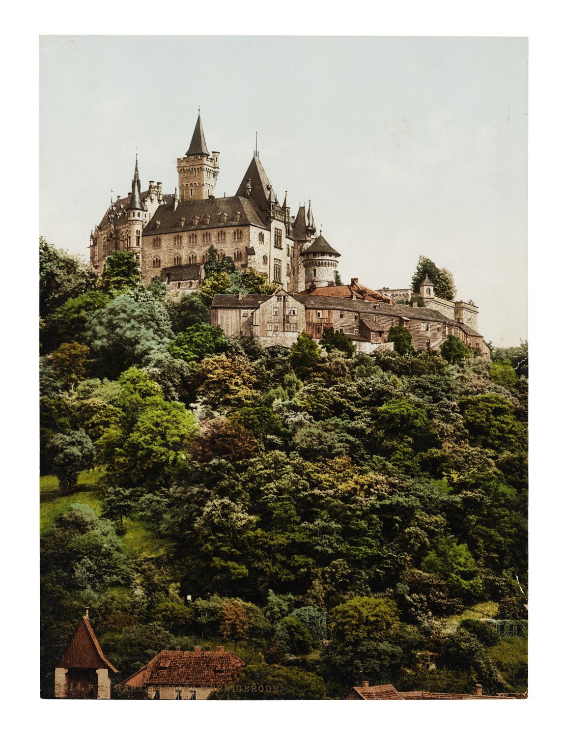 A colour photograph of a castle atop a treed hillside overlooking a village