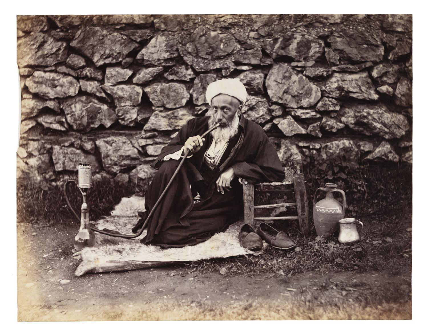A seated man with a bear smokes a hookah by a rock wall.
