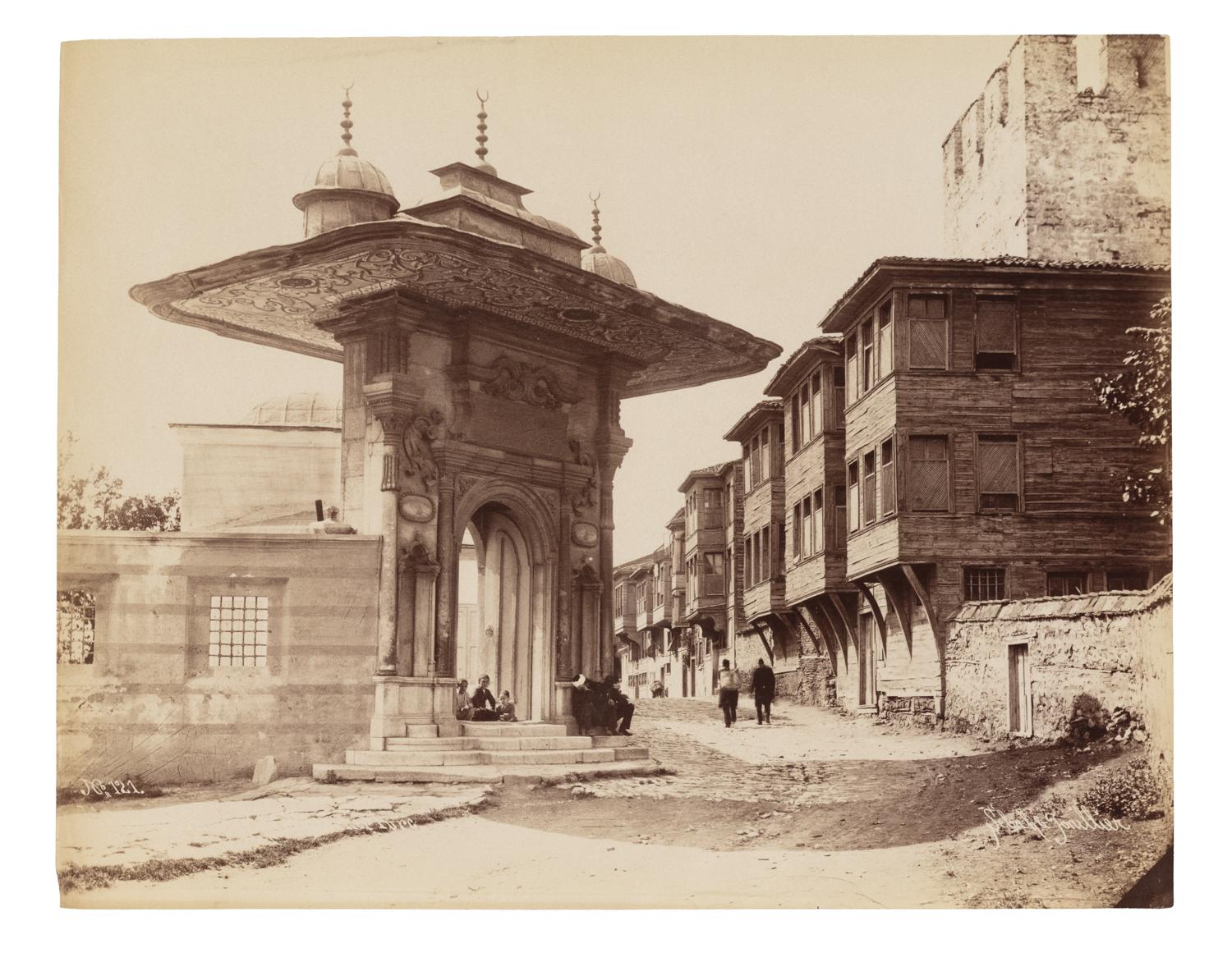 An ornate gate on a street with people and wooden structures on the right.