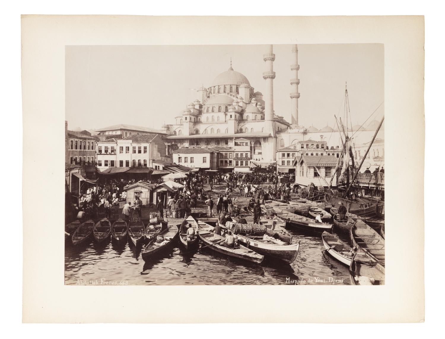 The photograph captures a bustling waterfront scene in Istanbul, centered around the Yeni Cami (New Mosque), a prominent Ottoman imperial mosque located in the Eminönü district near the Golden Horn.

In the foreground, the quay is crowded with small wooden rowboats and fishing vessels, some filled with goods, possibly fish or textiles. People are actively loading, unloading, or navigating the crowded shoreline, indicating a lively commercial hub or market area.

Behind the waterfront, a densely packed collection of Ottoman-era buildings leads the eye upward toward the monumental Yeni Mosque, with its grand central dome, semi-domes, and twin minarets rising into the sky. The mosque dominates the skyline and serves as both a religious and architectural focal point.

The photograph is sepia-toned and mounted on a white border, typical of late 19th-century albumen prints, and bears the studio mark: “Abdullah Frères, 663” in the bottom left, and
“Mosquée de Yeni-Djami” in the bottom right.