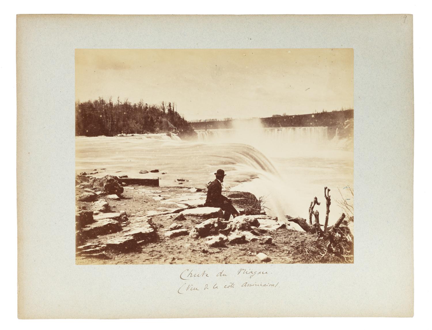 The photograph captures a serene yet powerful view of Niagara Falls, taken from the American side. The central figure in the image is a man in a dark suit and bowler hat, seated on a rock near the edge of the water. He faces the cascading falls, absorbed in the natural spectacle unfolding before him.

The waterfall is rendered in a soft blur, evoking the immense motion and mist of the crashing water. A long exposure technique has likely been used, lending a ghostly, ethereal quality to the rushing water. Spray rises from the falls, catching the light and creating a hazy, almost dreamlike effect.

The surrounding landscape is rugged and natural: rocky shoreline in the foreground, leafless branches to the right, and a tree-covered ridge in the distance on the left. The horizon is marked by the lip of the falls and low-lying structures or trees barely visible through the mist.