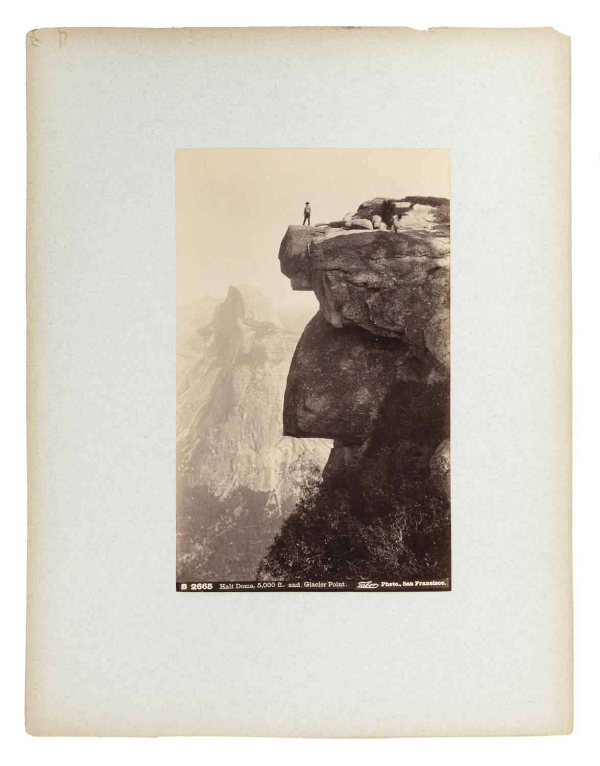 The composition showcases the awe-inspiring cliffs of Glacier Point, with a single figure standing boldly on the edge of a massive granite outcrop. Dressed in dark clothing and a hat, the person gazes out over the expansive Yosemite Valley, dwarfed by the immensity of the surrounding terrain.

In the background, the iconic Half Dome rises majestically, its sheer face glowing faintly in the distance. The vertical format of the photograph accentuates the dramatic elevation and vertical drop of the cliff, evoking a sense of both wonder and vertigo.

To the right of the figure, another person is crouched, possibly preparing camera equipment—suggesting a moment of exploration or early landscape documentation. The photo powerfully conveys themes of human scale against nature’s grandeur, a hallmark of early Western landscape photography.