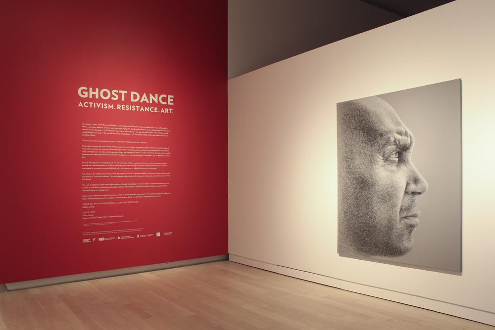 On the left, there is a red wall with white text that reads 'Ghost Dance. Activism. Resistance. Art.' On the right, a white wall with a photograph of a man's profile.