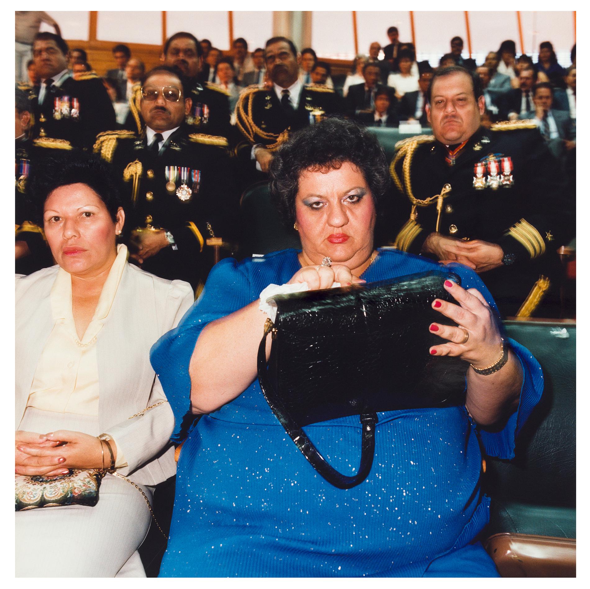 Seated in an auditorium, a woman in a blue dress looks directly at the camera; behind her sit military personnel