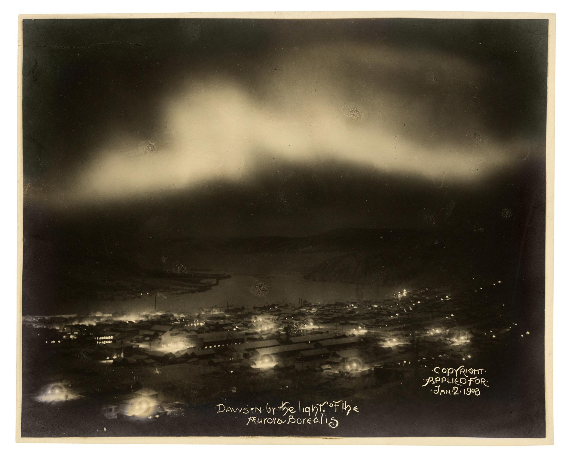 A black and white photograph overlooking a small town at night with lights and a band of aurora borealis in the sky