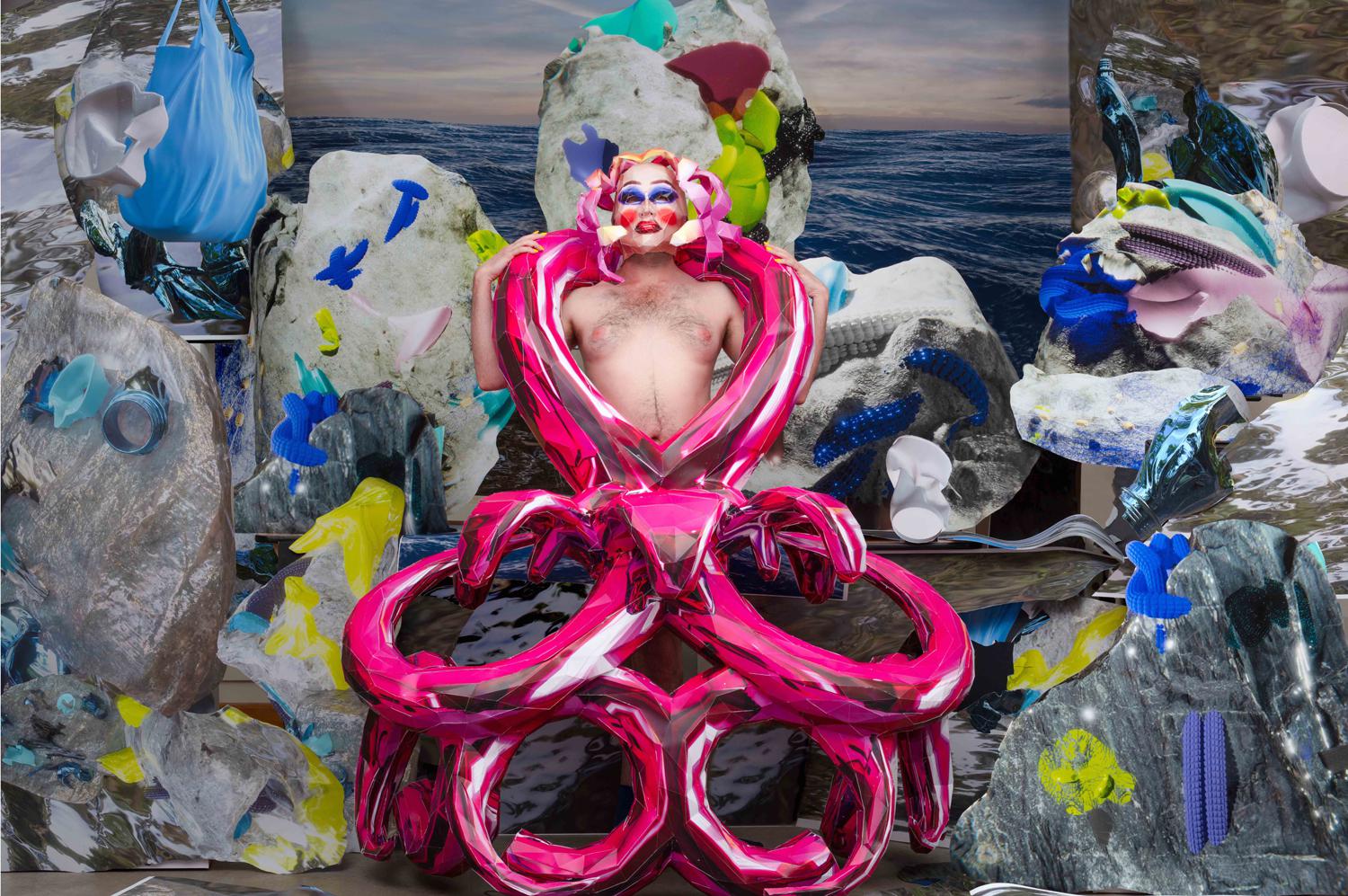 male figure with makeup stands shirtless shrouded in pink loops around his body and colourful shapes behind him. Image has cartoon like graphics.