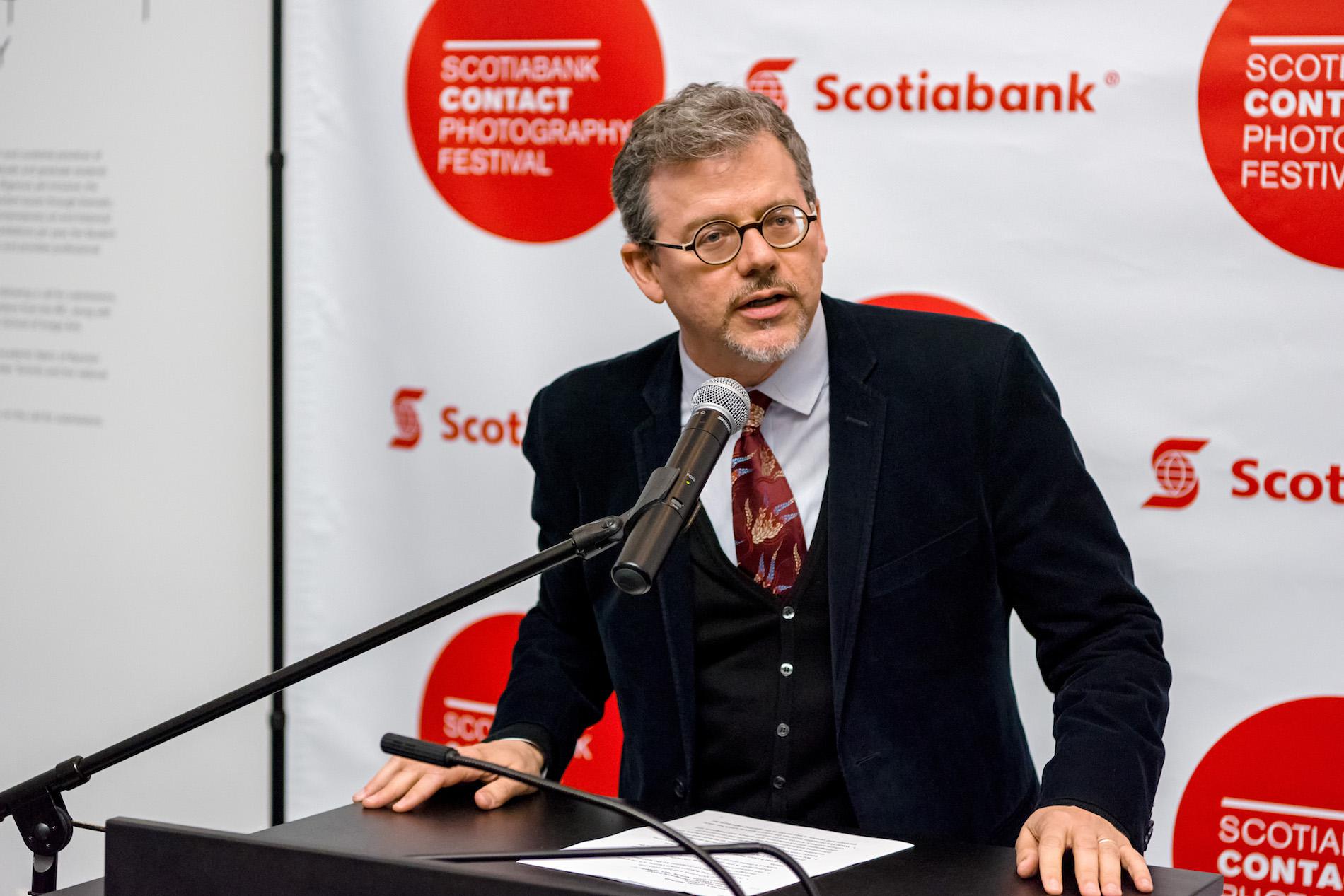 Man with circular glasses speaks into a microphone at a podium, in front of a backdrop that reads "Scotiabank Contact Photography Festival"