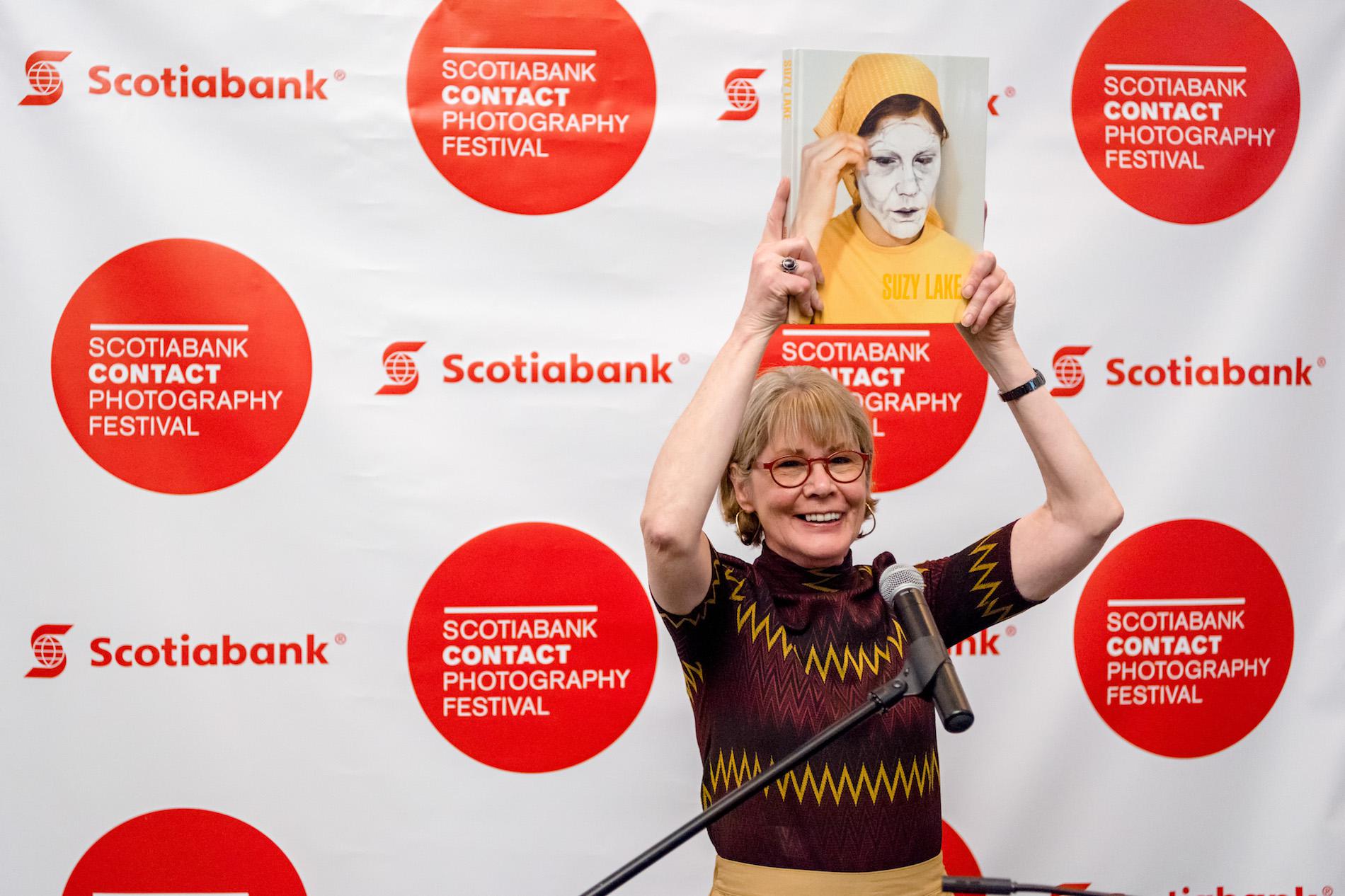 Artist Suzy Lake holds her book above her head and smiles. Backdrop reads 'Scotiabank Contact Photography Festival'