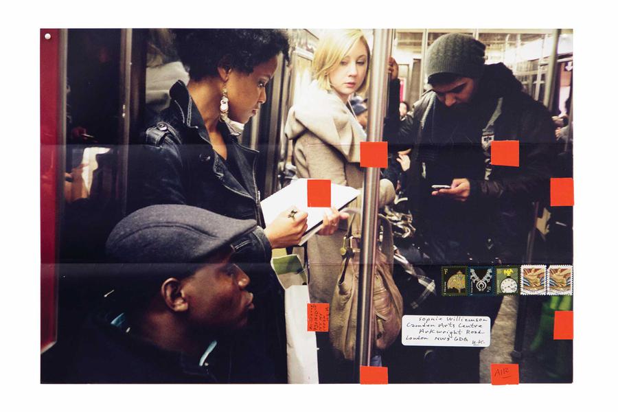 A group of four people stand near each other on the subway, one writing in a notebook; postage stamps, an address label and red stickers are pasted on top