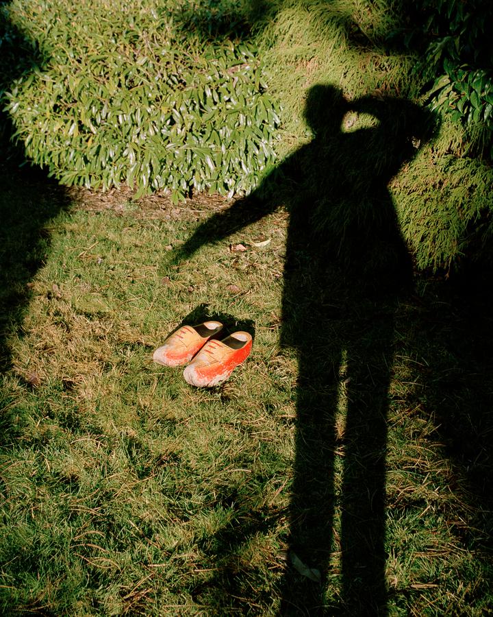 self-portrait of persons shadow on grass, orange clogs to the left of them