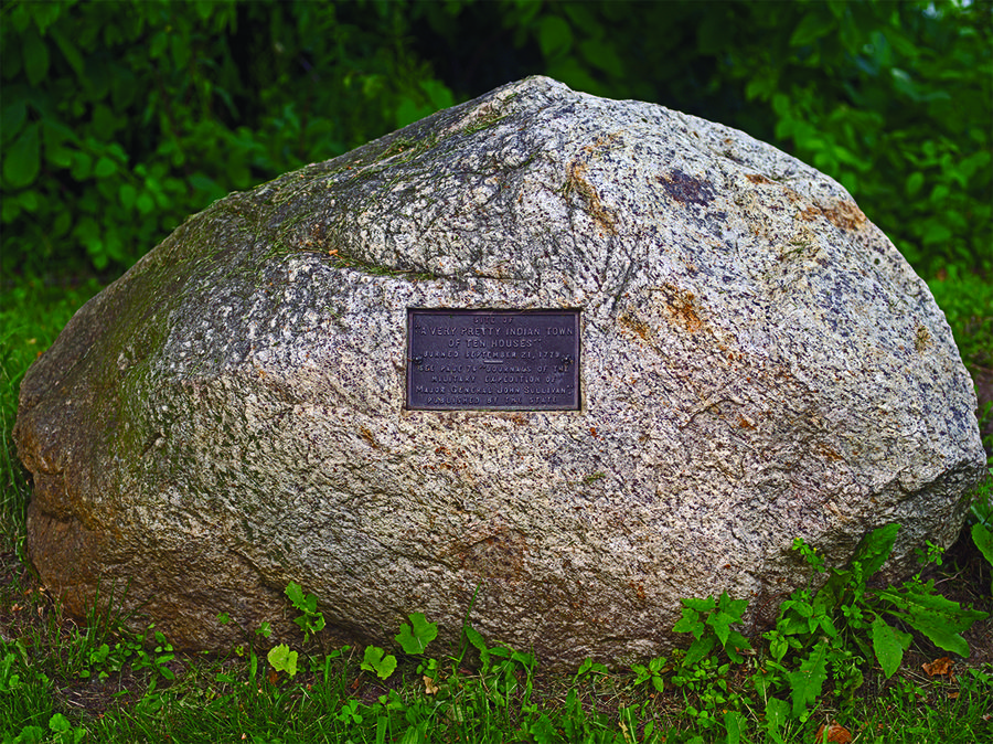 A large rock with a bronze historical plaque embedded into it is surrounded by grass
