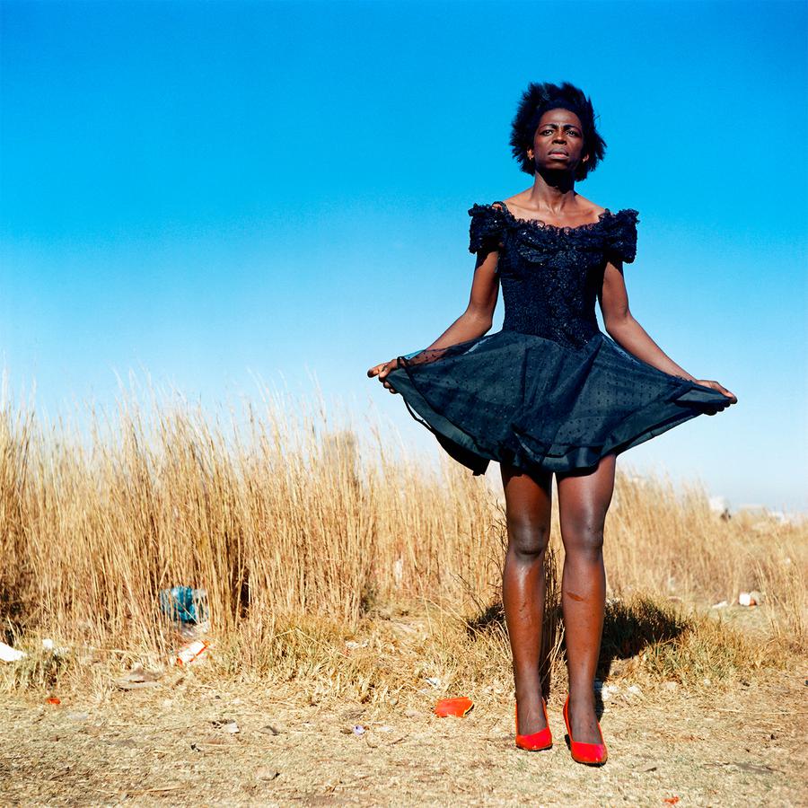 A individual stands on the edge of a field, in red shoes and a black dress.
