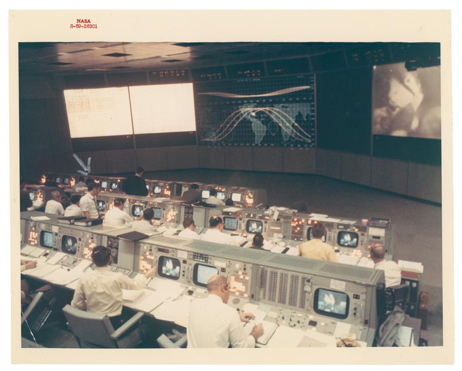 NASA control room with employees sitting in front of old computer screens and at the front of the room is a large screen with a map of the Earth showing the jet stream