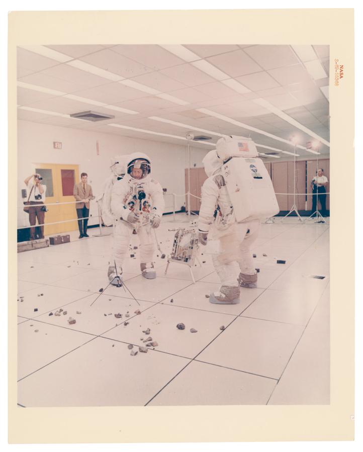 Two astronauts in space suits inside an office space practicing picking up rocks