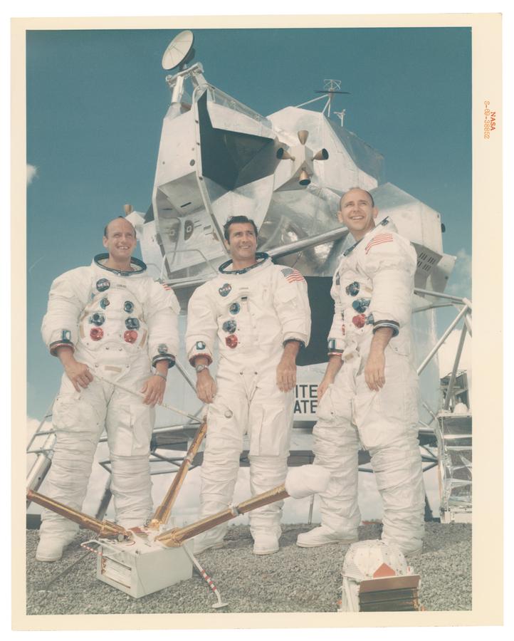 Three NASA Astronauts stand in front of a space ship with their helmets off smiling