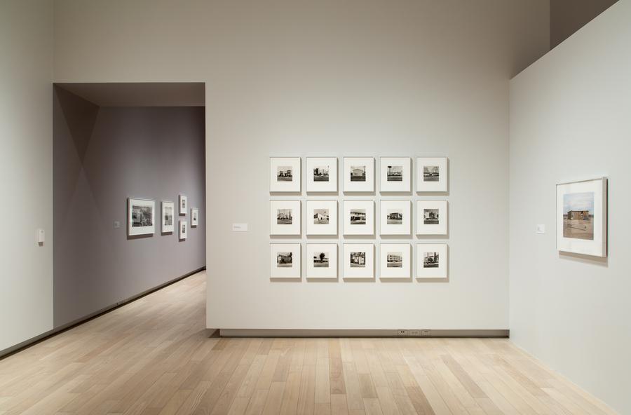 Grid of framed photographs on a grey wall, to the left an opening to another gallery space