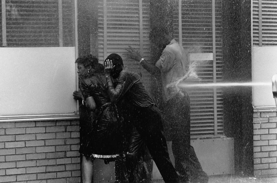 Firemen blast black protestors with water, protestors duck against a brick wall