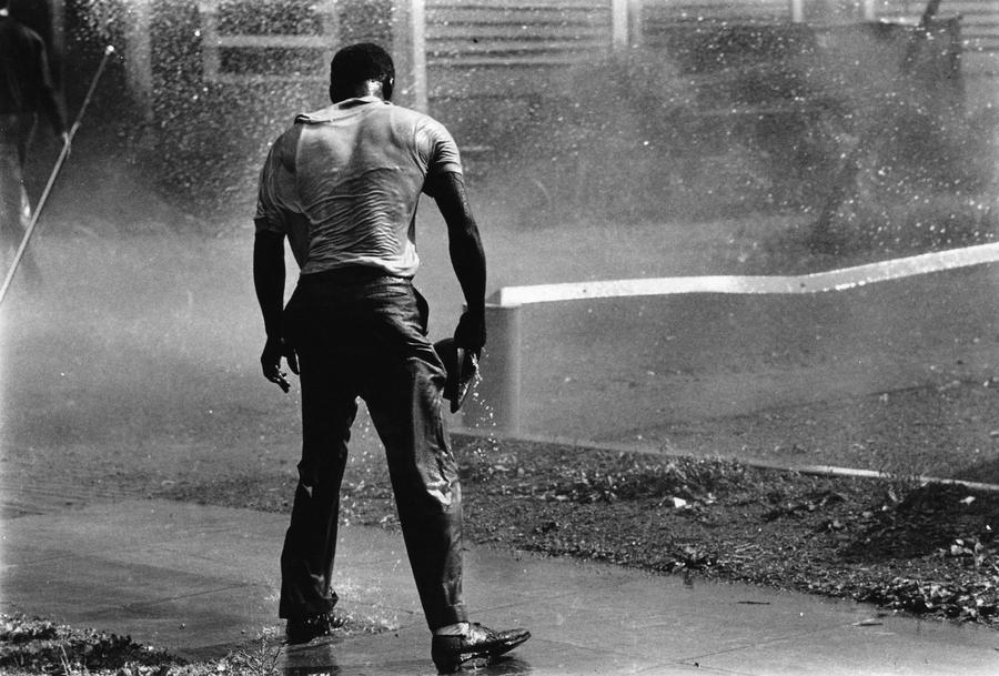 A man in a tshirt and pants stands on the sidewalk soaking wet