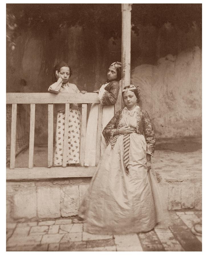 Three young women pose together on a verandah in dresses.