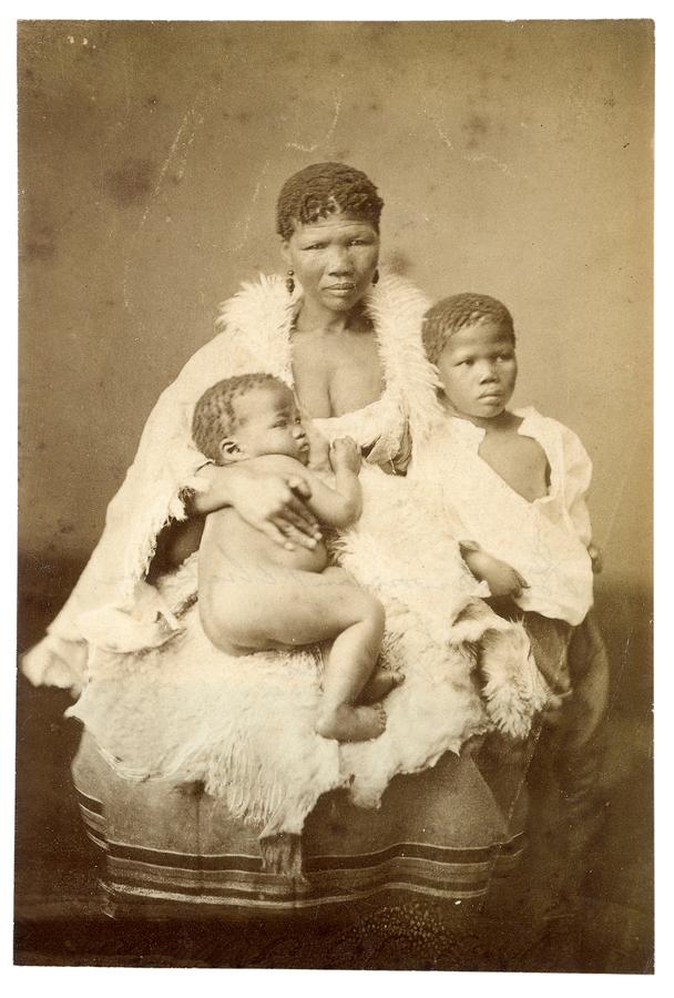 This early photograph shows a woman seated 
and clothed in a cloak, with two young children on her lap