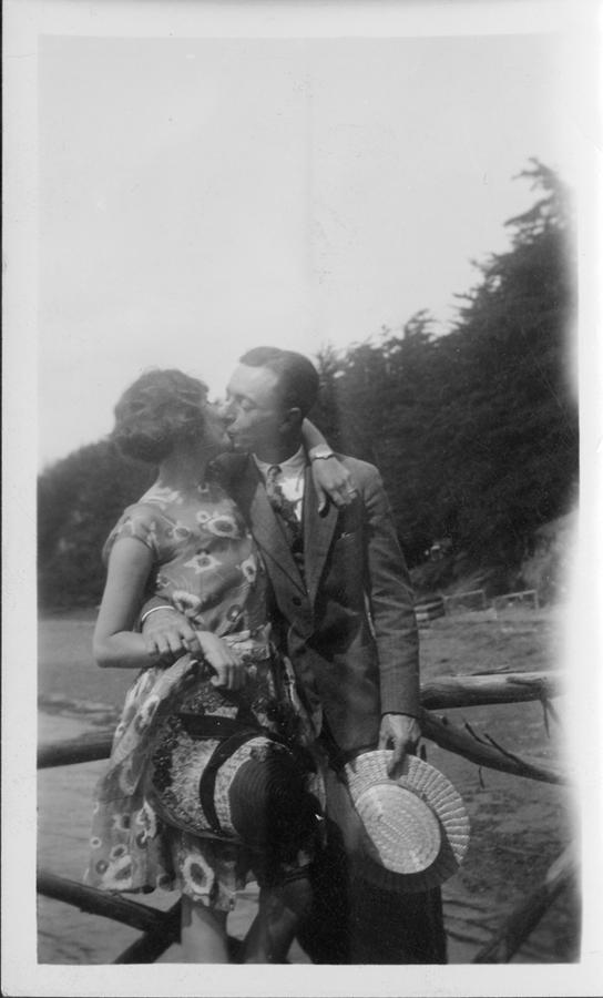 A man and woman, each holding a hat, kiss, with a view of a beach and trees in the background.