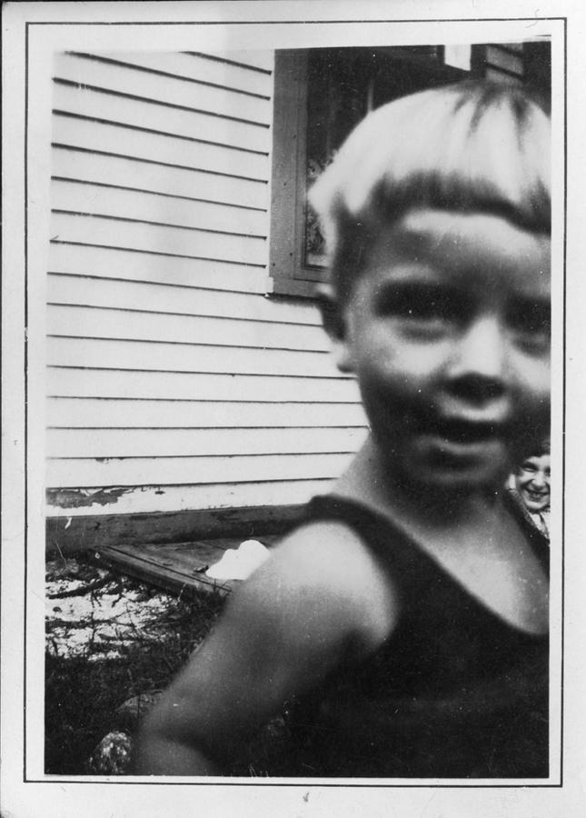 A black and white photograph shows a young boy with blonde hair and wearing a sleeveless top standing close to the camera lens and pictured out of focus. Behind him is a nondescript building, in focus.