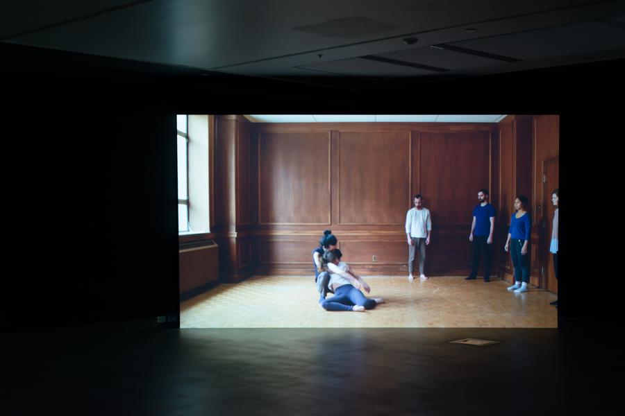 Video still of a man and woman holding each other in the centre of a room, while four people stand along the walls