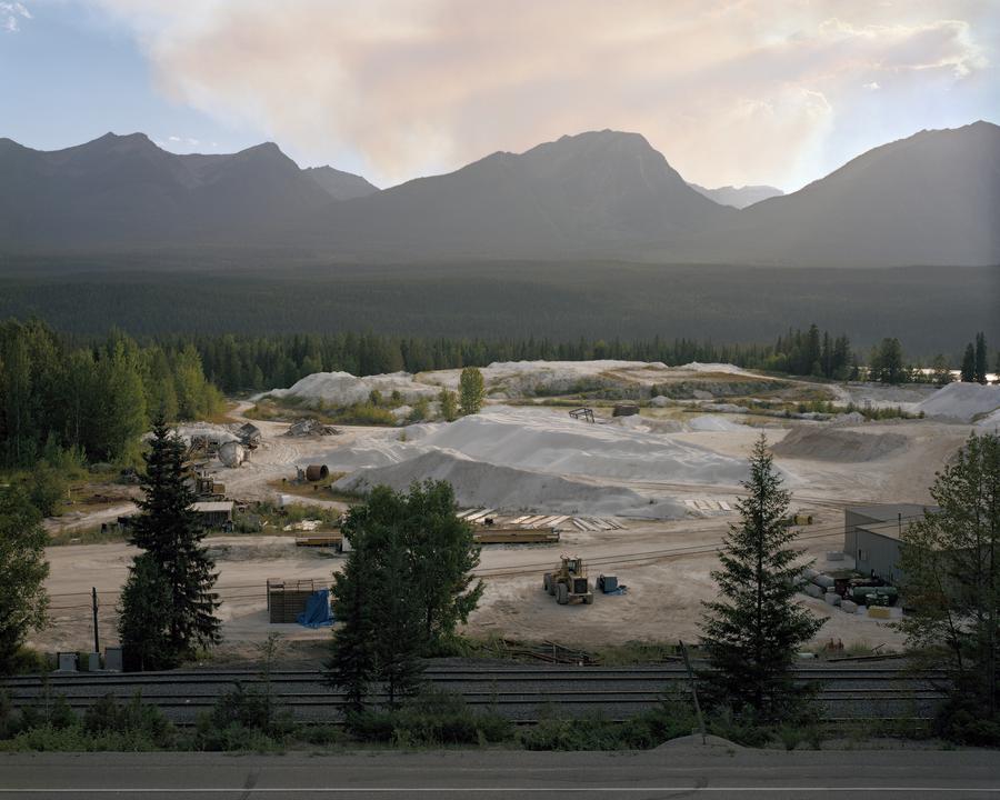 A plume of smoke rises from behind a mountain range. In the foreground, piles of sand and construction equipment